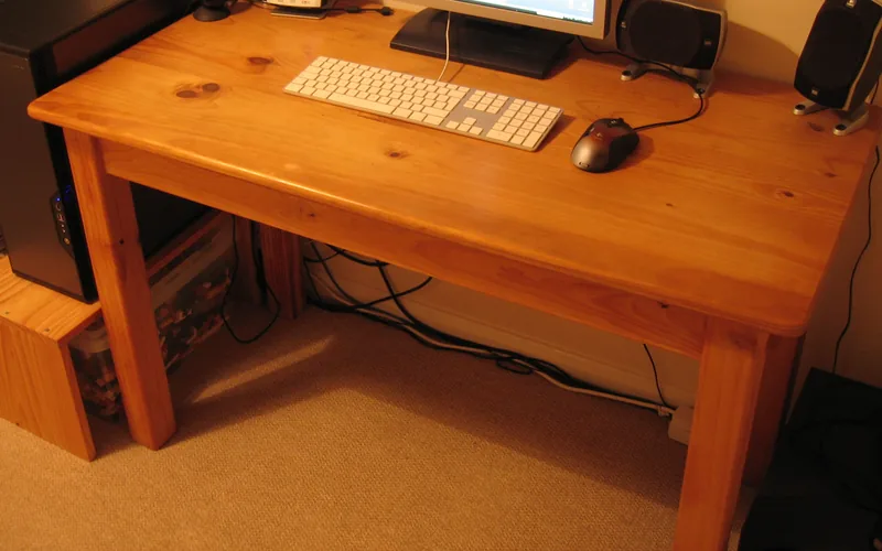 Mesa de pino barnizado con la peana de un monitor, un teclado blanco, un ratón y altavoces; debajo se aprecian los cables
