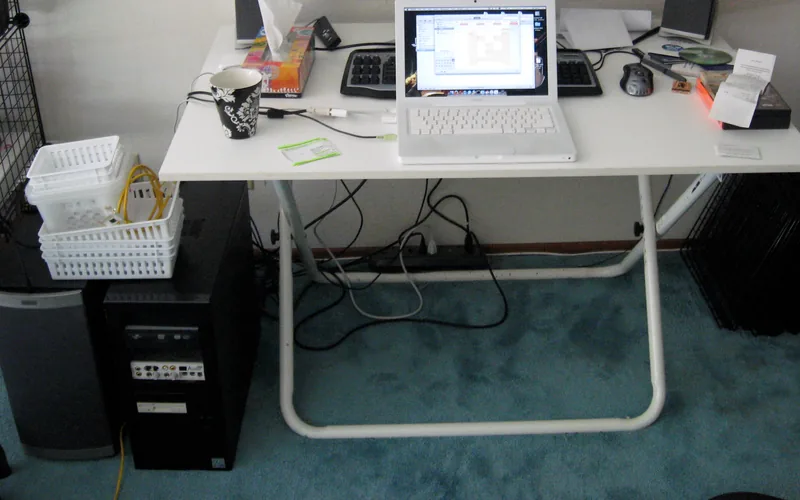 Mesa plegable blanca con un portátil abierto, un teclado y una taza; bajo el tablero cuelgan cables hasta una regleta en el suelo