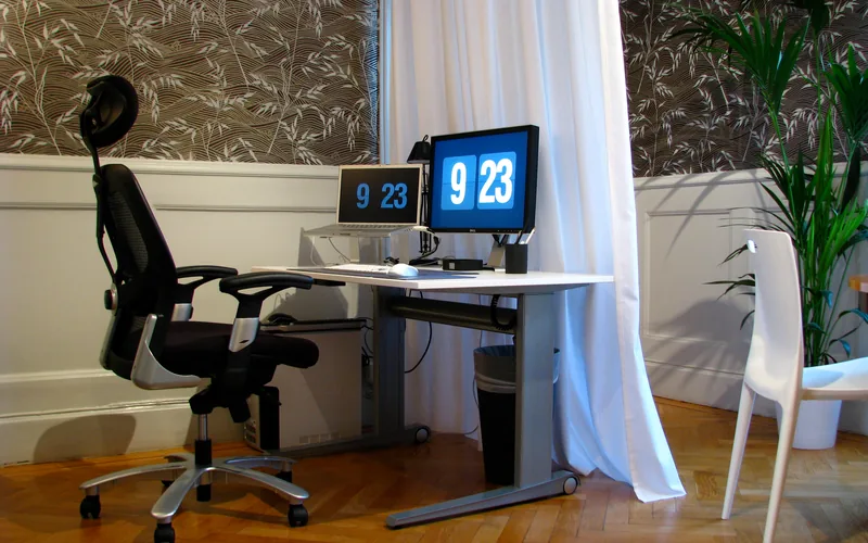 Mesa blanca de altura regulable con un portátil elevado y un monitor que muestran un reloj; a la izquierda, una silla de oficina negra